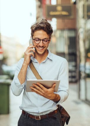 Ein junger Mann mit Brille im weißen Hemd telefoniert und schaut dabei auf ein Tablet