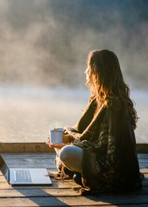 Eine Frau sitzt entspannt mit einer Kaffeetasse vor einem Laptop an einem nebelverhangenen See am Morgen