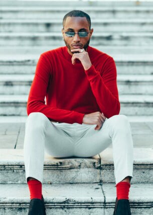 Ein Mann in weißen Hosen und einem roten Kaschmirpullover sitzt auf einer weißen Marmirtreppe