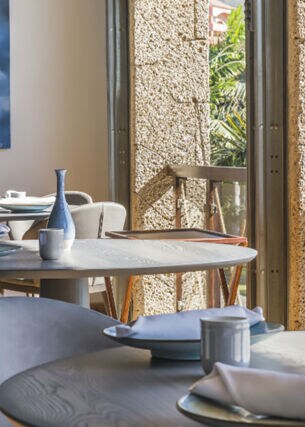 Innenraum eines luftigen, hellen Restaurants mit gedeckten Tischen an einer Fensterfront mit Blick auf Palmen