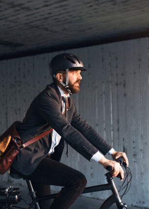 Seitenansicht einer Person im Anzug, mit Umhängetasche und Helm, die auf einem Fahrrad fährt