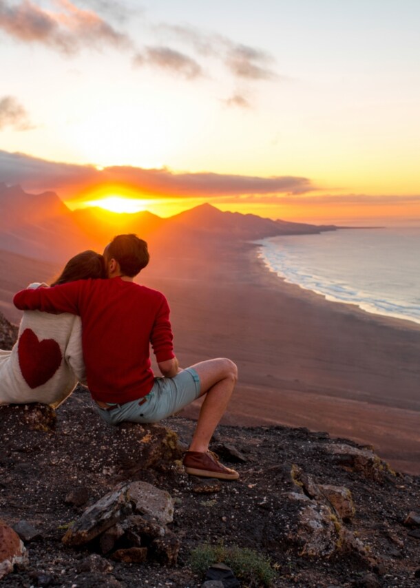 Zwei Personen sitzen mit dem Rücken zur Kamera auf einem Felshang und Blicken in den Sonnenuntergang über den Bergen, rechts im Bild das Meer