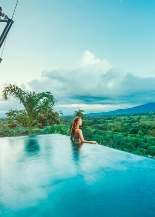 Profilansicht einer Frau, die aus einem Infinity-Pool über eine tropische Hügellandschaft schaut