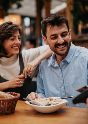 Eine Frau mit braunen Haaren und ein Mann mit Bart sitzen im Restaurant und lächeln, während er dem Kellner seine Kreditkarte zum bezahlen reicht