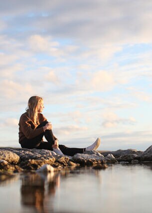 Junge Frau mit blonden langen Haaren sitzt auf Felsen und schaut übers Meer