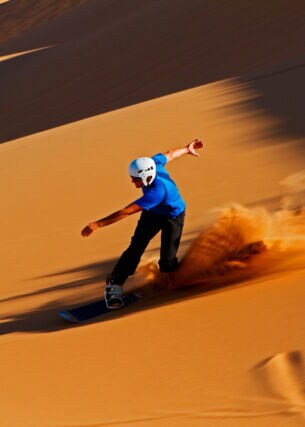 Ein Sandboarder mit Helm fährt eine rötliche Sanddüne hinunter