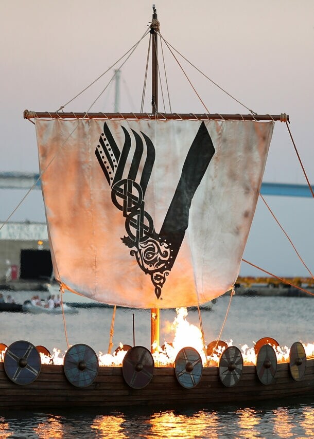 Aufnahme eines brennenden Wikingerschiffs auf dem Meer mit einem Segel, das das Logo der „Vikings“-Serie zeigt