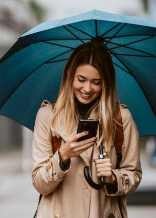 Eine junge Frau auf einer Straße schaut lächelnd auf ihr Smartphone unter einem Regenschirm
