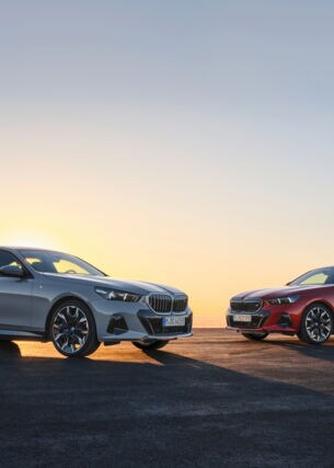 Zwei BMW i5, einmal in Silber, einmal in Rot, die von hinten von der Sonne angestrahlt werden.