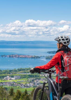 Rückansicht einer Frau mit einem Fahrrad vor dem Panorama des Bodensees