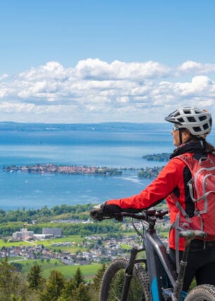 Rückansicht einer Frau mit einem Fahrrad vor dem Panorama des Bodensees
