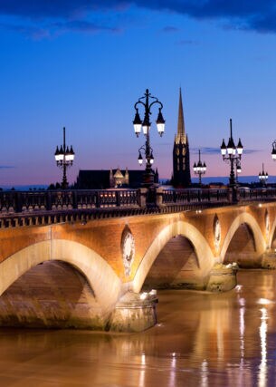 Eine alte Steinbrücke mit erleuchteten Straßenlaternen verläuft über einen Fluss in Bordeaux bei Nacht