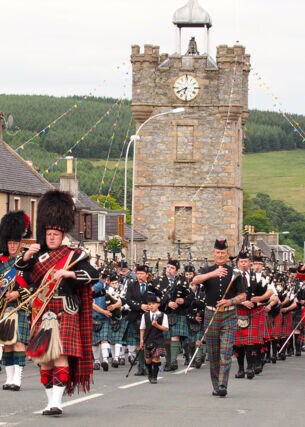 Diverse Menschen in traditionellen schottischen Outfits, die eine Straße entlanglaufen und Instrumente spielen.