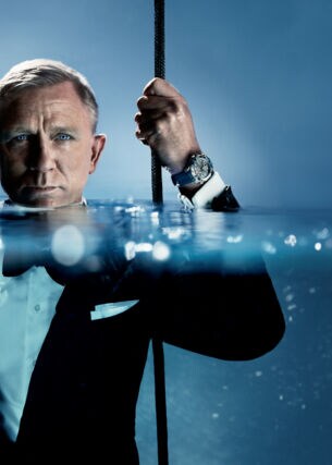 Porträt von Daniel Craig im schwarzen Smoking im Wasser, das ihm bis zum Hals steht; an seiner hochgehaltenen Hand trägt er eine Taucheruhr der Marke OMEGA