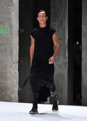 Designer Rick Owens in einem schwarzen, lässigen Kleid läuft in einer Industriehalle über einen Lauftsteg