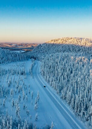 Luftaufnahme einer weiten Winterlandschaft mit schneebedeckten Wäldern, durch die ein einzelnes Auto über eine Straße durch den Schnee in der Abenddämmerung fährt
