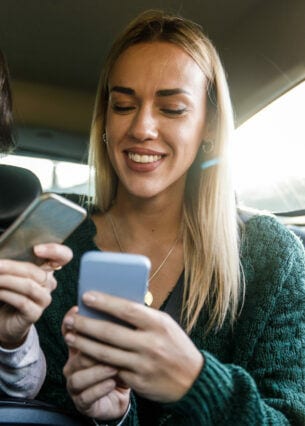 Zwei junge Frauen auf der Rückbank eines Autos schauen lächelnd auf ihre Smartphones