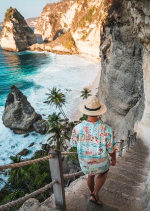 Rückansicht eines Mannes mit Strohhut und Hawaiihemd, der eine schmale Steintreppe an einer Felswand hinunter zu einer palmengesäumten Strandbucht geht