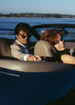 Schauspieler Pierce Brosnan mit Sonnenbrille sitzt neben einer Frau am Steuer eines Cabriolets
