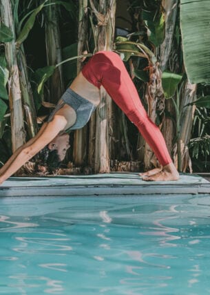 Eine junge Frau bei einer Yogaübung auf einer Matte an einem Pool, umgeben von Palmen