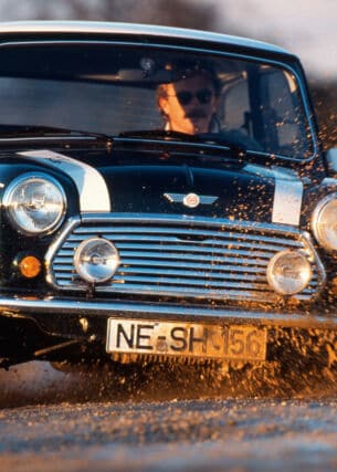 Ein Mann fährt in einem schwarzen Mini Cooper 1.3 mit Rallystreifen durch eine Schlammpfütze