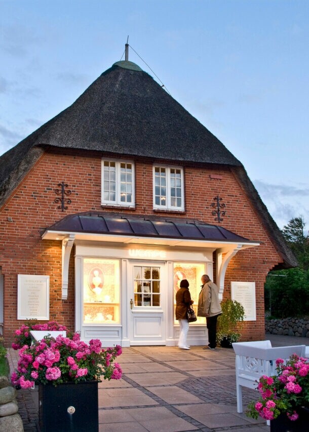 Zwei Personen stehen vor einem beleuchteten Schaufenster einer Luxusboutique in einem reetgedeckten Haus