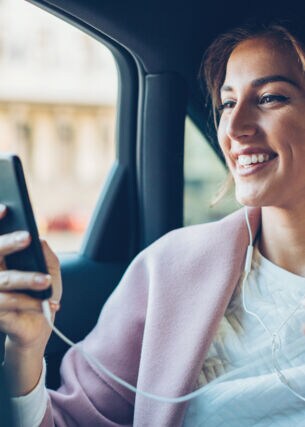 Eine junge Frau mit Kopfhörern auf dem Rücksitz eines Autos schaut lachend auf das Display ihres Smartphones.