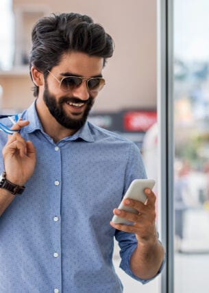 Ein junger Mann mit Vollbart, Sonnenbrille und Einkaufstüten in der Hand schaut in einer Einkaufsstraße lächelnd auf sein Smartphone.