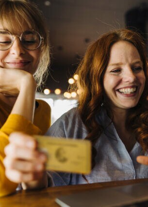 Zwei junge Frauen sitzen lächelnd mit einer Kreditkarte vor einem Laptop in einem Café.