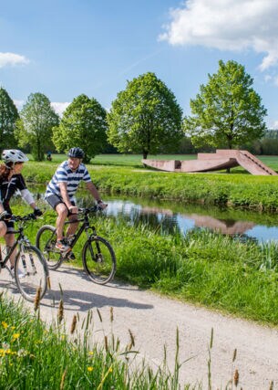 Zwei Personen auf Fahrrädern fahren auf einem Radweg durch eine grüne Landschaft entlang eines Flusses.