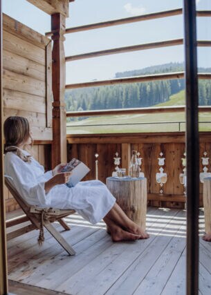 Eine Frau in weißem Bademantel sitzt mit einem Buch auf einer Terrasse eines Holzchalets mit Blick in die Berge