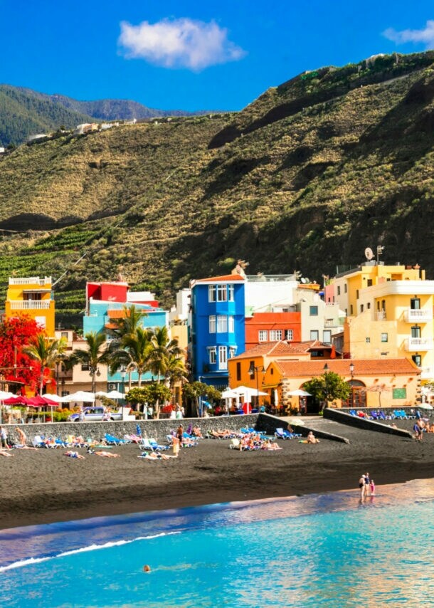 Schwarzer Sandstrand mit Badenden in einem Ort mit bunten Häusern vor Bergpanorama
