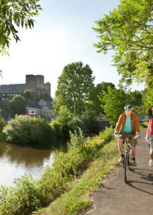 Ein älteres Paar fährt auf Fahrrädern auf einem asphaltierten Radweg entlang eines Flusses, im Hintergrund eine mittelalterliche Burgruine in einer Ortschaft