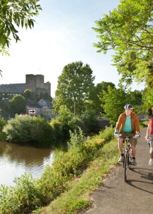 Ein älteres Paar fährt auf Fahrrädern auf einem asphaltierten Radweg entlang eines Flusses, im Hintergrund eine mittelalterliche Burgruine in einer Ortschaft