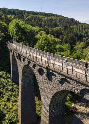 Drei Radfahrende fahren auf einem schmalen Weg auf einem Viadukt in einer hügeligen Waldlandschaft