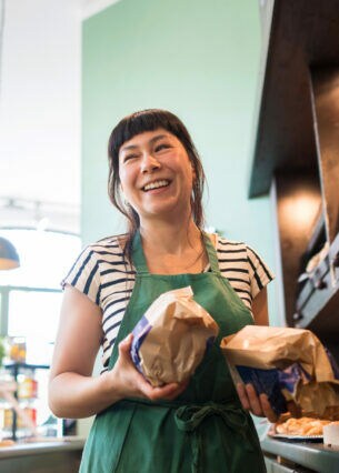 Eine Verkäuferin mit Backwaren in den Händen steht lachend hinter der Theke einer Bäckerei.