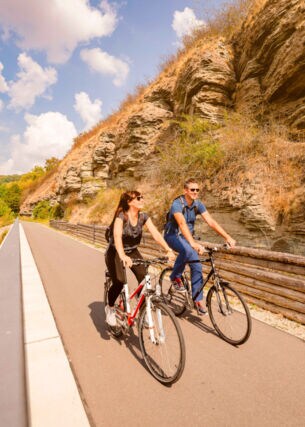 Zwei Personen fahren auf Fahrrädern auf einem asphaltierten Radweg vor einer Steinwand an einem Fluss entlang