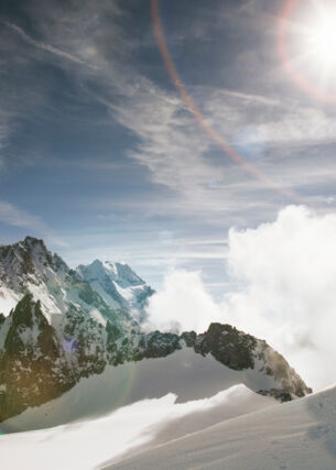 Zwei Personen gehen einen schneebedeckten Bergkamm entlang, im Hintergrund erhebt sich ein noch höheres Bergpanorama