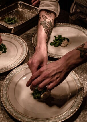Nahaufnahme von Händen beim Anrichten von Speisen auf großen, weißen Tellern in einer Restaurantküche.