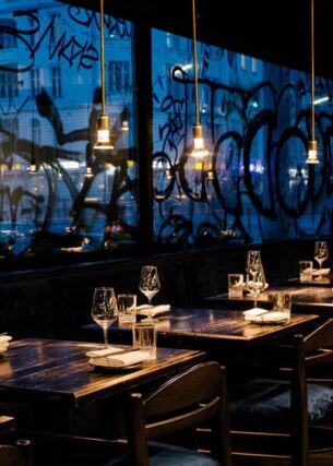 Beleuchtete, eingedeckte Holztische in einem modernen Restaurant vor einer Fensterfront mit Graffiti bei Nacht.