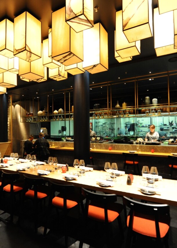 Ein langer, eingedeckter Tisch in einem modernen Restaurant im fernöstlichen Stil mit offener Küche bei gedimmter Beleuchtung.