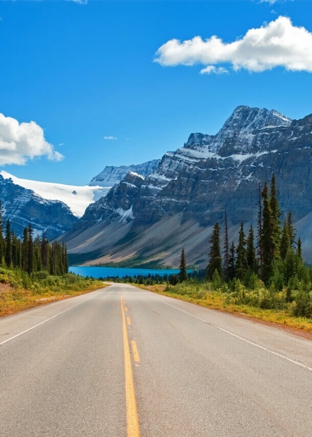 Ein Highway, umringt von Nadelbäumen, im Hintergrund schneebedeckte Berge und ein türkisblauer See.