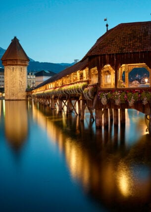Eine überdachte und beleuchtete Holzbrücke über einem Fluss, ein Wasserturm und eine Stadt und Berge im Hintergrund.