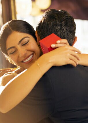 Eine Frau mit einer roten Geschenkbox in den Händen umarmt glücklich lächelnd einen Mann auf einer Sitzbank in einem Restaurant.