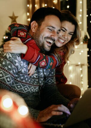 Ein lachendes Paar in Winterpullovern sitzt aneinander gekuschelt auf einem Sofa im Wohnzimmer vor einem Laptop.