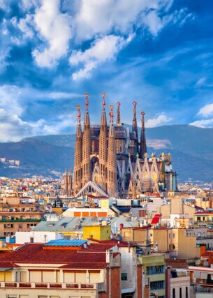 Stadtpanorama von Barcelona mit der emporragenden Sagrada Familia vor einem Gebirgszug im Hintergrund.