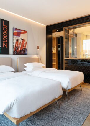 Elegantes Hotelzimmer im modernen Design mit zwei Betten und offenes Badezimmer hinter einer Glasscheibe.