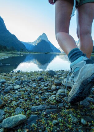 Zwei Beine einer Person in Wanderschuhen an einem See vor Bergpanorama.