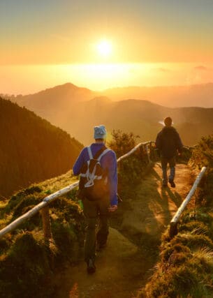 Rückansicht zweier gehender Personen auf einem Weg entlang eines Bergkamms inmitten einer grünen Gebirgslandschaft bei Sonnenuntergang.