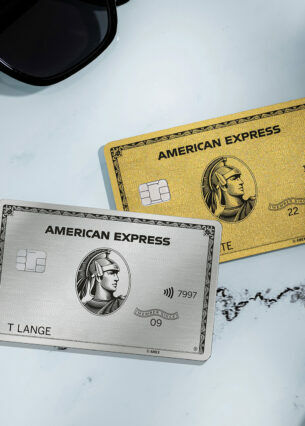 Eine silberne und eine goldene Kreditkarte von American Express liegen auf einem weißen Marmortisch neben einer schwarzen Sonnenbrille, einer Kaffeetasse und einem Teller mit Gebäck.
